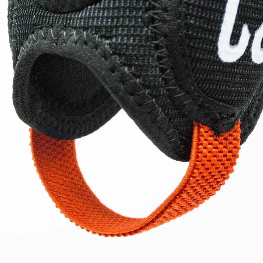 Close-up of the CORE Ankle Skate Protection Guards in black textured fabric, featuring an orange strap looped through it, perfect for scootering or BMXing. The edges are reinforced with stitching and part of the CORE logo is visible. The focus is on the strap and fabric details, crafted for those seeking durable ankle protection.