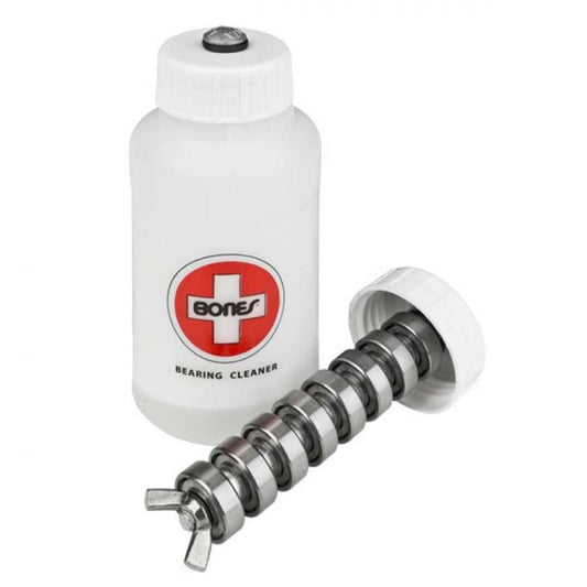 A white cylindrical container labeled with the product name "Bones Bearing Cleaning Unit," made from fluorinated high-density polyethylene, features a red cross logo associated with the Bones brand. Next to it is an opened lid and a metal rod specifically designed for the precise cleaning of skateboard bearings.