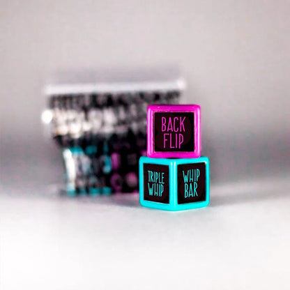 Two vibrant dice from the Trick Dice Expert Scooter Set are stacked with flair: one pink showcasing the trick "Back Flip" and another blue featuring "Triple Whip" and "Whip Bar." Crafted for advanced dice maneuvers, these capture the bold essence of professional rider Terry Price. In the blurred background, additional dice offer exciting trick combinations.