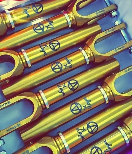 Close-up of seven gold scooter forks, including the Apex Sig Jesse Bayes Gold Quantum Threadless edition, arranged in a row. Each is labeled "Apex." They rest on a reflective surface, highlighting the shiny finish and detailed engravings on each fork.