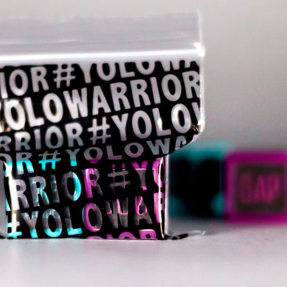 A close-up of the glossy black and silver Trick Dice Advanced Scooter Set box, featuring the repeated "#YOLOWARRIOR" text, hints at the thrill of a new trick challenge. The background, blurred with unrecognizable pink and blue objects, captures the adventurous spirit associated with Trick Dice's pro rider Terry Price.