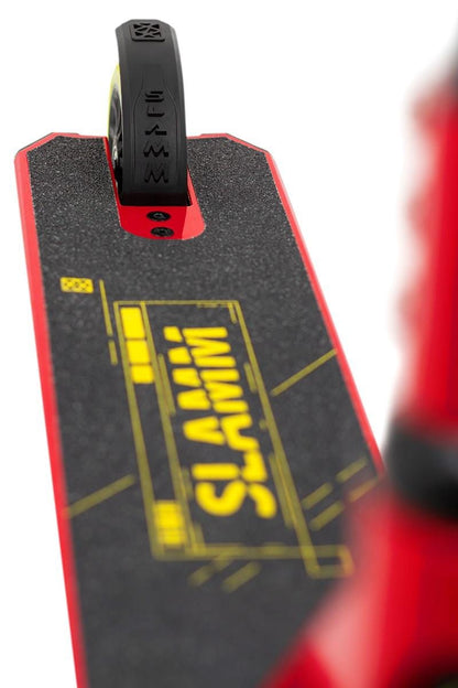 Close-up of the Slamm Classic Mini Complete Stunt Scooter - Red deck, featuring black grip tape with "SLAMM" in yellow. The rear freestyle scootering wheel prominently shows the embossed "SLAMM" brand name on the tire, all set against a bright white background.