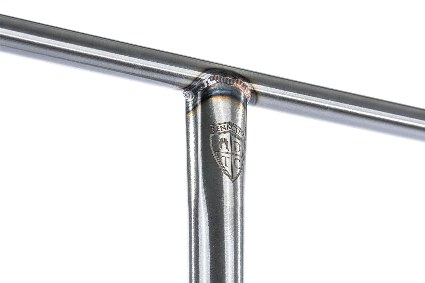 Close-up of a metallic T-bar joint from the Ethic DTC Tenacity V2 Steel HIC Stunt Scooter, showcasing precise welding and a small engraved shield emblem. Its lightweight, polished surface reflects light, emphasizing a smooth, industrial finish akin to Ethic craftsmanship.