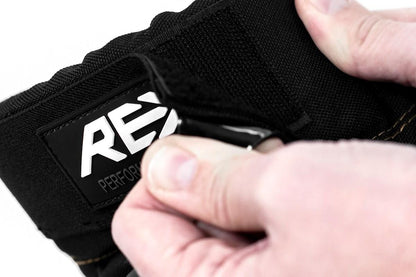 Close-up of hands adjusting a black strap with the visible "REKD" label, suggesting impact resistance suitable for action sports. The material appears durable, showcasing clearly defined stitches. The softly blurred background keeps the focus on the hands and strap, reminiscent of REKD Ramp Knee Skate Protection Pads in Black/Khaki.