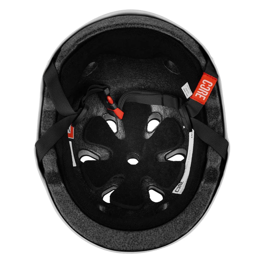 An overhead view of a white CORE Action Sports Skate/Scooter Helmet reveals the inside padding, ventilation holes, and adjustable black straps with red accents. The interior lining of the helmet is visible, along with tags labeled "CORE.