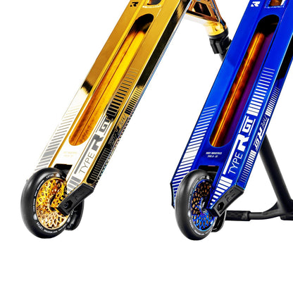 Close-up of two stunt scooters with intricate designs and vibrant colors. One, a gold sleek model, stands out with its detailed graphics and small wheels enhanced by glittering rims from Root Industries. The other is the Blu-Ray variant of the Root Industries Type R GT Complete Stunt Scooter, showcasing a striking blue finish.