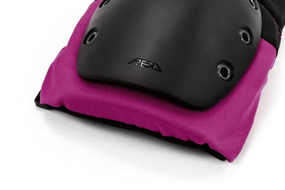 Close-up of the REKD Ramp Knee Skate Protection Pads in Black/Pink, highlighting the black protective gear with pink fabric edging and multiple rivets. These pads are designed for action sports and feature a smooth, curved surface to ensure optimal impact resistance.