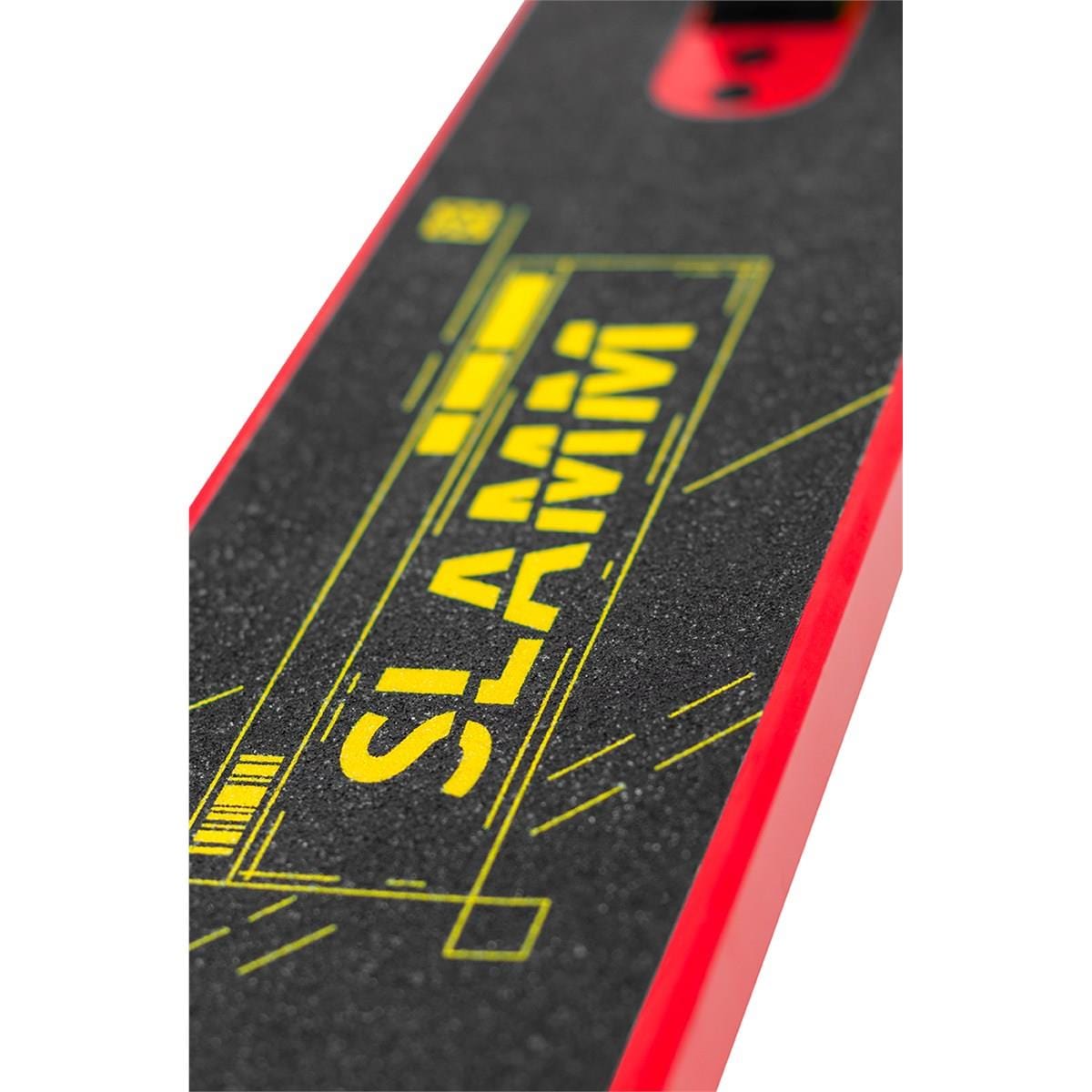 A close-up of the Slamm Classic Mini Complete Stunt Scooter reveals its skateboard-like deck covered with black grip tape featuring the brand name "SLAMM" printed in vibrant yellow, capturing the bold energy typical of freestyle scootering. The edges boast a striking red color, with glimpses of an intricate graphic design visible through the grip tape.