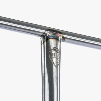 Close-up of a raw chrome silver T-joint with a welded connection, reminiscent of the Ethic DTC Tenacity Steel Oversized HIC Stunt Scooter Bars' robust construction. A small emblem with "TENACITY" and a shield design is visible on the vertical bar, showcasing the durability of the Ethic brand's Tenacity V2. The background is plain white.