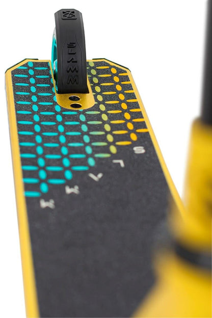 Close-up of the Slamm Assault Complete Stunt Scooter - Gold, showcasing its alloy deck adorned with a vibrant grid design in blue, green, and yellow dots. The deck features a textured surface for enhanced grip. A small rear wheel and 110mm aluminum wheels are prominently seen at the top of the deck.