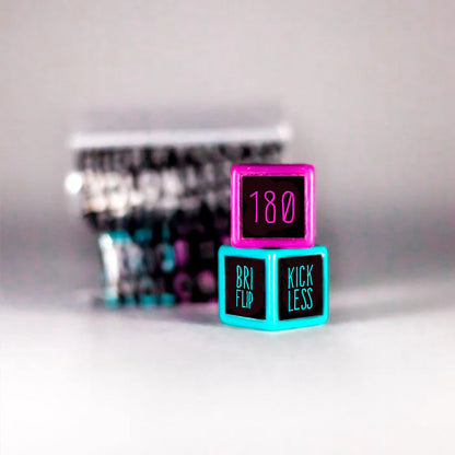 Two vibrant dice from the Trick Dice Advanced Scooter Set, ideal for a trick challenge, are meticulously stacked. The top die is pink and displays "180," while the bottom one is blue and features "BRI FLIP" alongside "KICKLESS." A softly blurred background enhances the focus on these Trick Dice products, evoking the expertise of professional rider Terry Price.