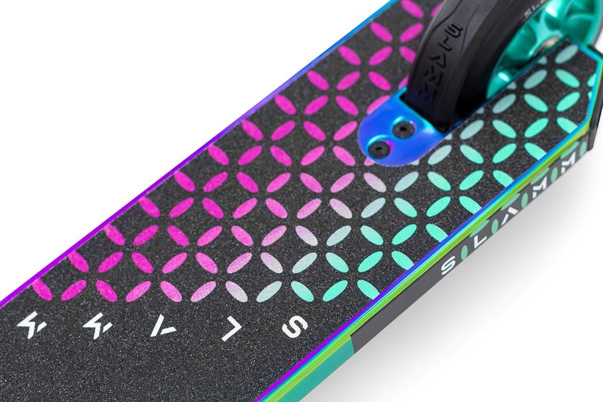 Close-up of the Slamm Assault Complete Stunt Scooter - Neochrome alloy deck, showcasing a vibrant pattern of overlapping circles in pink, blue, and purple hues. The surface features a textured grip with prominent brand lettering, accompanied by the partially visible 110mm aluminum wheels.