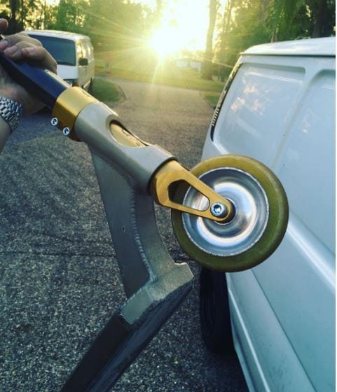 Close-up of a person holding a yellow polyurethane caster wheel attached to a metal bracket, resembling the sleek design of the Apex Sig Jesse Bayes Gold Quantum Threadless Scooter Fork. The background reveals a street with a white van and sunlight streaming through the trees.