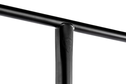 Close-up of the Ethic DTC Tenacity V2 Steel Oversized HIC Stunt Scooter T-Bars in black, showcasing a T-shaped joint. The smooth metal surface is adorned with an engraved shield logo, embodying the essence of stunt scooter craftsmanship. The lighting accentuates its sleek design and lightweight construction.