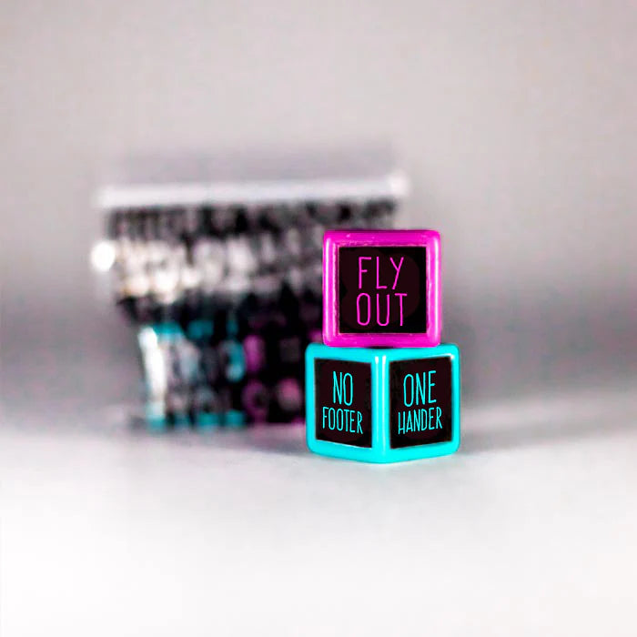 Two vibrant dice from the Trick Dice Beginner Scooter Set are stacked, featuring a top pink die that reads "Fly Out" and a bottom blue die displaying "No Footer" and "One Hander." This Trick Dice challenge is perfect for any pro rider ready to take on daring dice tricks with blurred backdrops of similar dice in anticipation.