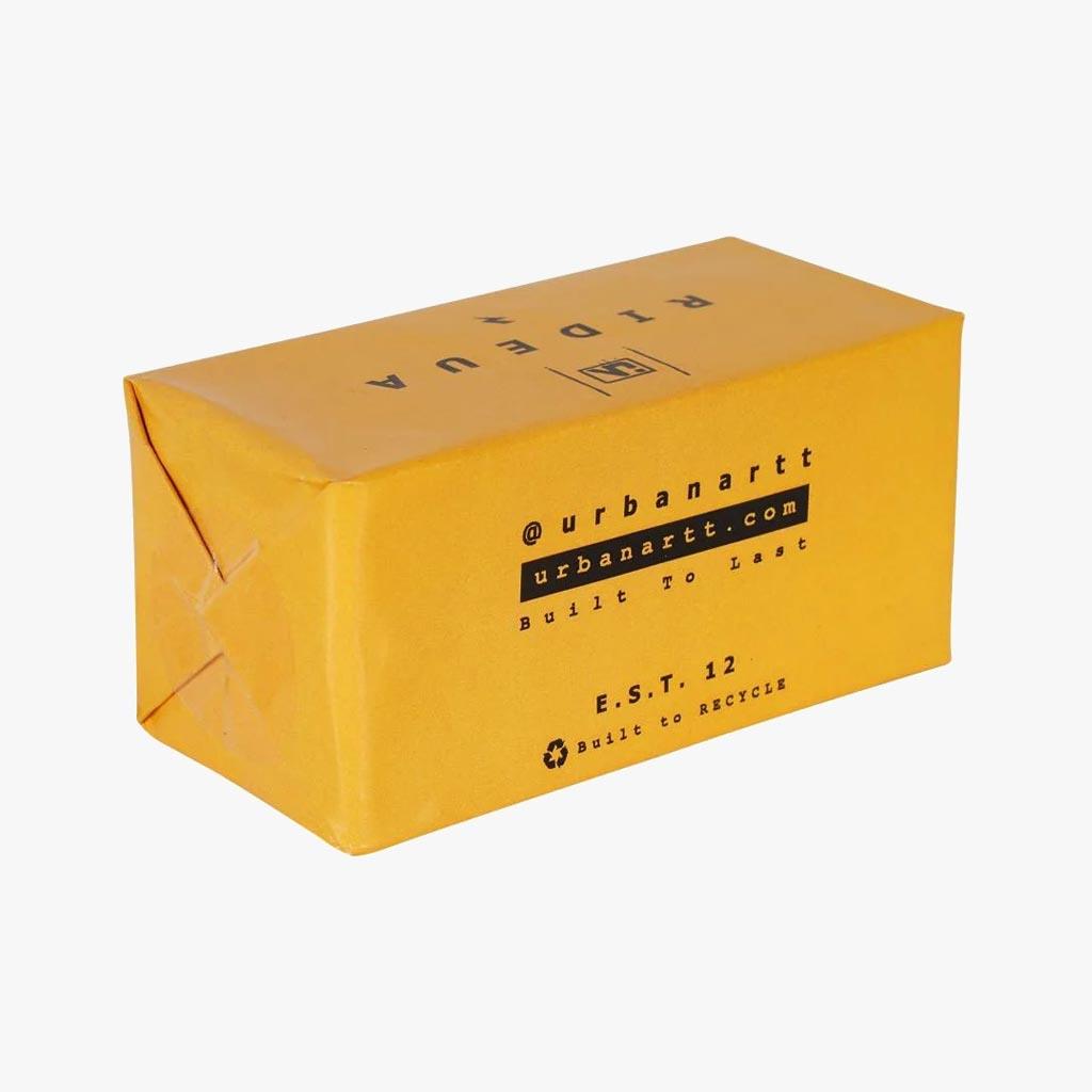 The UrbanArtt Butter Block Wax - Yellow comes in a yellow rectangular cardboard box adorned with the stylish imprint of "@urbanartt," "urbanartt.com," "Built to Last," and "E.S.T. 12." Designed for smooth performance, its folded edges emphasize a minimalist aesthetic that ensures durability and recyclability.