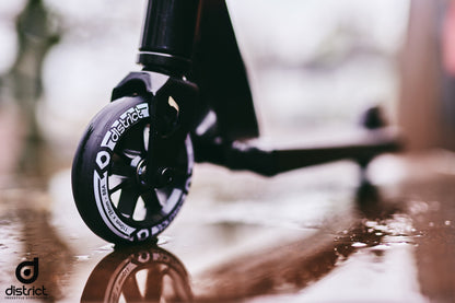 A close-up shot showcases the wheel of a District Titus Complete Stunt Scooter - Black on a glistening, wet surface, capturing reflections of light. The word "District" is prominently displayed on the wheel, part of this beginner-friendly scooter. The blurred background accentuates the premium quality of its components and reveals a glimpse of the sleek scooter frame.