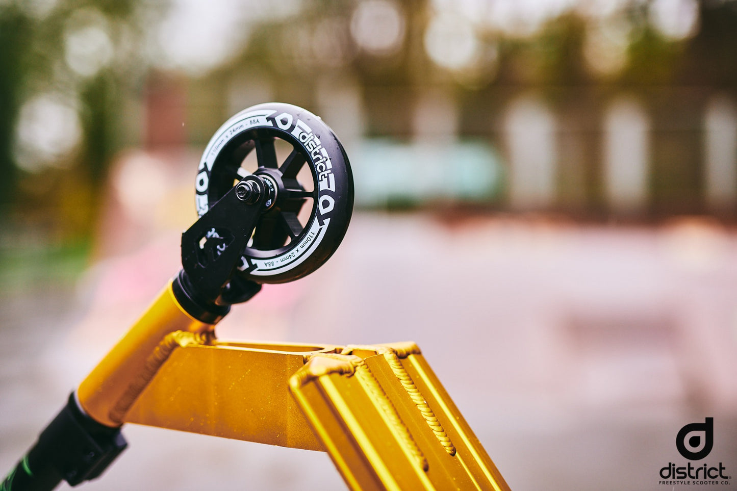 Close-up of a gold and black scooter from the District brand, ideal for beginner-level riders, with a prominently branded black wheel. The background is blurred, showcasing an outdoor skate park. The District Titus Complete Stunt Scooter logo is proudly displayed in the bottom right corner.