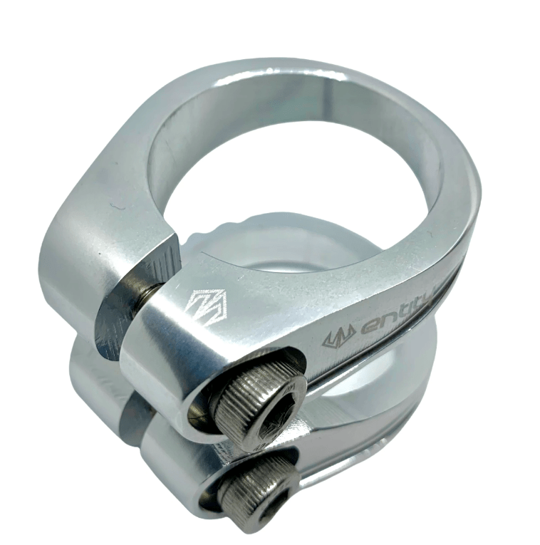 The Entity Exile 2 Bolt Oversized Stunt Scooter Clamp in raw silver is crafted using advanced 5 axis CNC machining technology. It features a bolt fastening mechanism with an engraved logo and is designed to be lightweight. This clamp boasts a sleek, polished finish and is shown against a reflective white background.