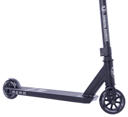 The Longway Adam Complete Stunt Scooter - Black is shown in a close-up, highlighting its lightweight black frame, two small wheels, and durable metal construction. The deck has a textured surface with "ADAM" printed on it, and the handlebar exhibits branding and design features perfect for beginner riders.