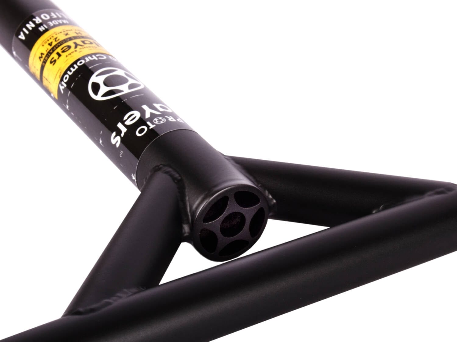 The close-up reveals a Proto Baby Slayer Classic Pro Steel SCS Stunt Scooter Bar in flat black, highlighting the chromoly steel construction and textured finish. The focus is on the Y-shaped joint and central tube, featuring a circular design with multiple holes. A yellow and black Proto logo sticker is prominently displayed on the top part, evoking the design of a Classic Stunt Scooter Bar.