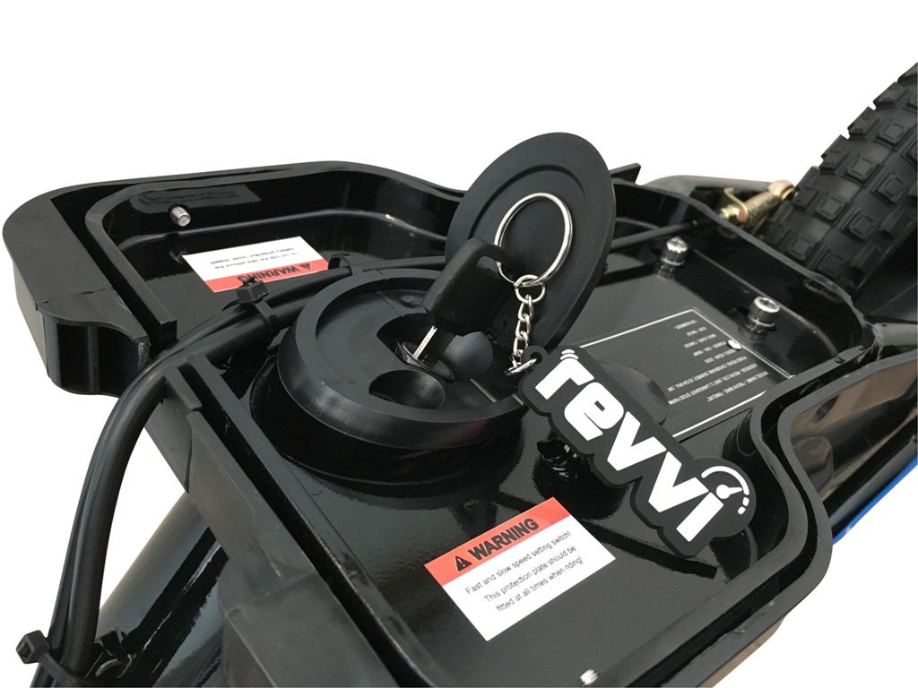 A close-up view shows a black key from the Revvi 16" Plus Kids Electric Balance Bike inserted into the battery compartment, featuring a protective covering and a visible warning label. In the background, part of the bike tire suggests its durable design characteristic of kids' electric bikes.