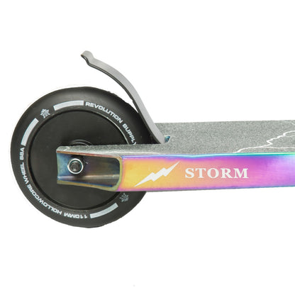 A close-up of the Revolution Storm Complete Stunt Scooter's rear wheel and deck highlights the black hollow core wheels adorned with white text. The Neochrome Storm Deck showcases a rainbow metallic finish, featuring "STORM" and a lightning bolt logo, with a metal brake positioned above the wheel.